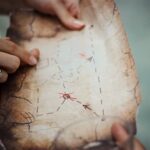 A pair of hands holds a hand drawn map, while a third hand points to a square on it.